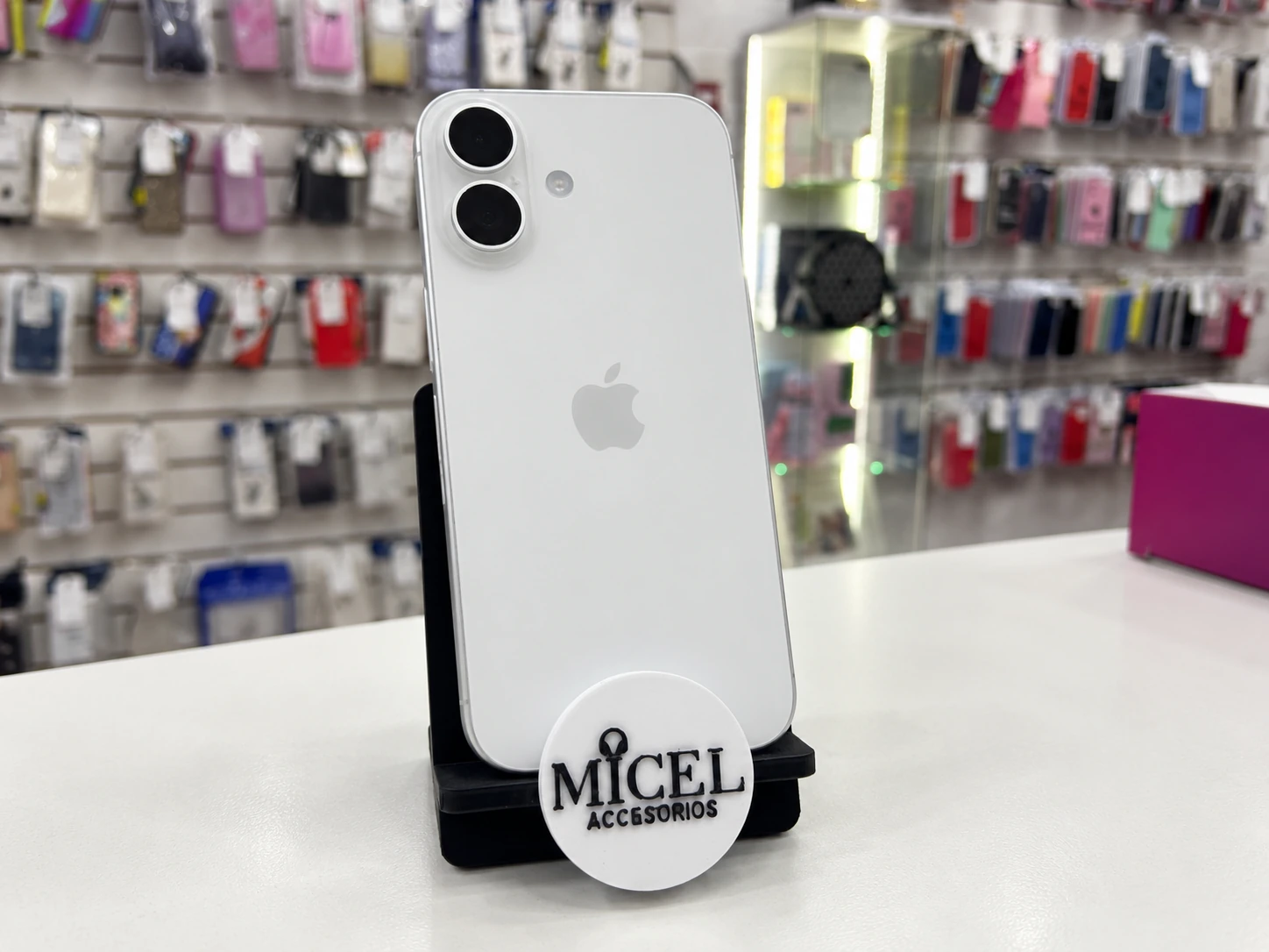 Imagen ilustrativa de iPhone blanco en soporte negro MICEL, con fondo de tienda.